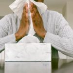 Woman in grey sweater sits down and holds tissue up to her nose.