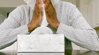 Woman in grey sweater sits down and holds tissue up to her nose.