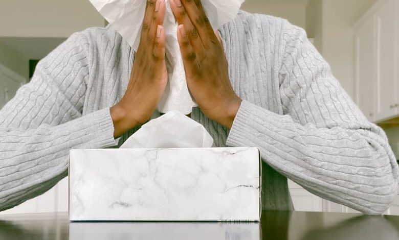 Woman in grey sweater sits down and holds tissue up to her nose.