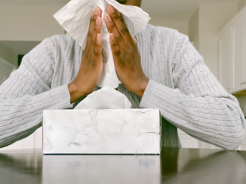 Woman in grey sweater sits down and holds tissue up to her nose.