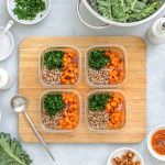 Quinoa, kale and roasted sweet potato lunch boxes are standing on a kitchen table with ingredients for cooking, overhead view, fall healthy take-away lunch recipe concept