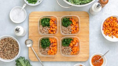 Quinoa, kale and roasted sweet potato lunch boxes are standing on a kitchen table with ingredients for cooking, overhead view, fall healthy take-away lunch recipe concept