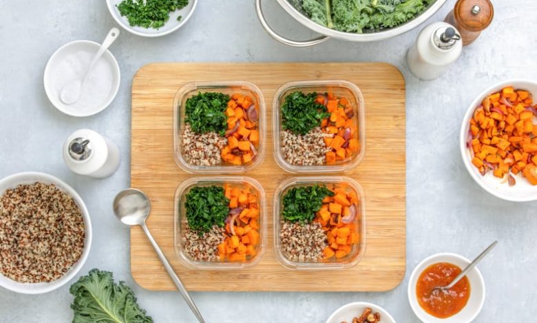 Quinoa, kale and roasted sweet potato lunch boxes are standing on a kitchen table with ingredients for cooking, overhead view, fall healthy take-away lunch recipe concept