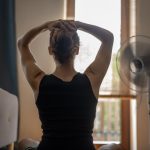 A young woman with her hands behind her head in front of a fan.