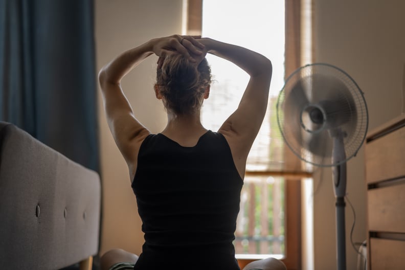 A young woman with her hands behind her head in front of a fan.