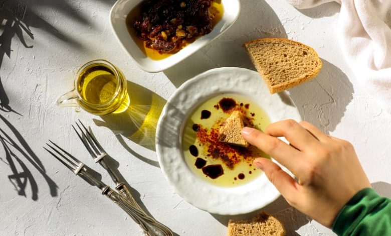 A woman dips a torn piece of hand made artisan bread in extra virgin olive oil on table top shot.