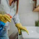 Young woman holding bottle of blue disinfectant while wiping down bathroom sink and wearing yellow rubber gloves.