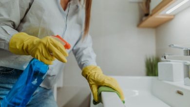 Young woman holding bottle of blue disinfectant while wiping down bathroom sink and wearing yellow rubber gloves.