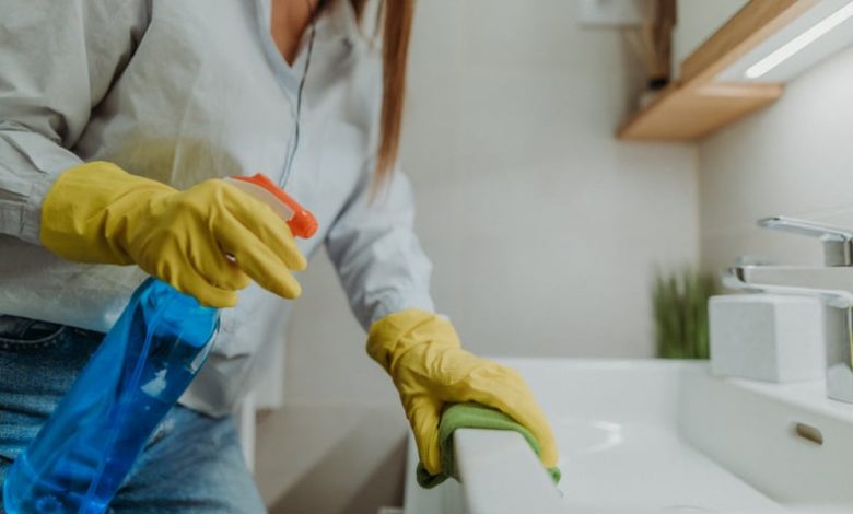 Young woman holding bottle of blue disinfectant while wiping down bathroom sink and wearing yellow rubber gloves.