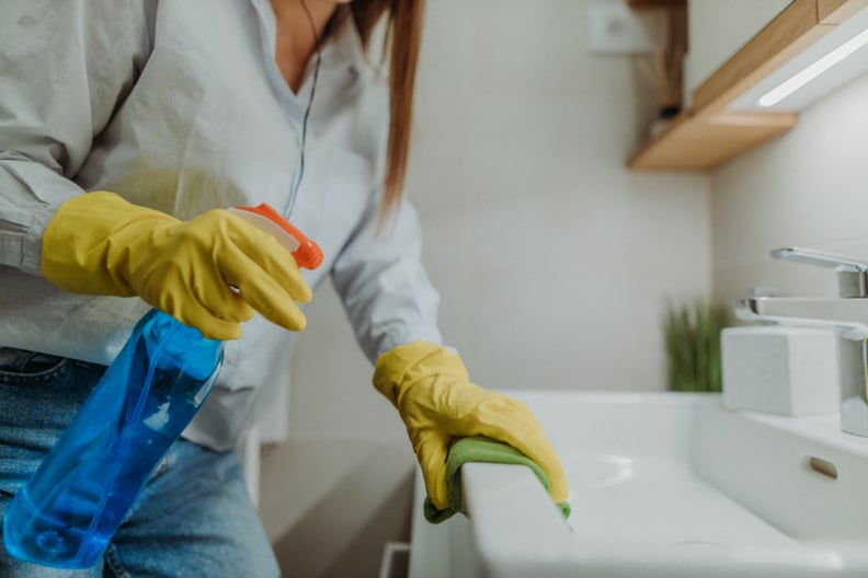 Young woman holding bottle of blue disinfectant while wiping down bathroom sink and wearing yellow rubber gloves.