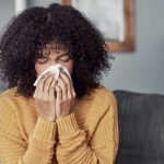 Shot of a young woman blowing her nose with a tissue at home, wondering if she can get her flu shot while sick.