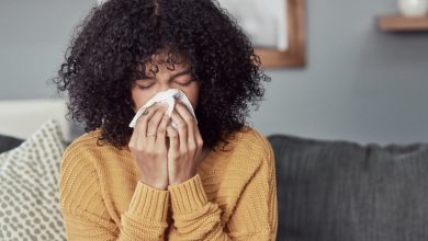 Shot of a young woman blowing her nose with a tissue at home, wondering if she can get her flu shot while sick.