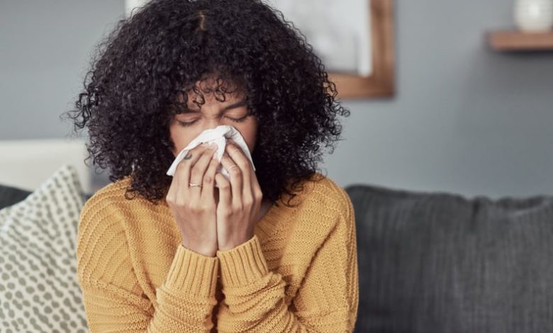 Shot of a young woman blowing her nose with a tissue at home, wondering if she can get her flu shot while sick.