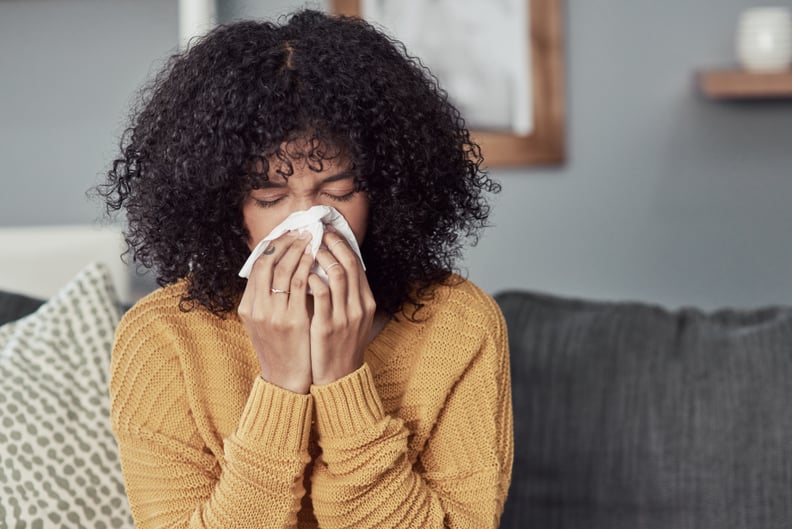 Shot of a young woman blowing her nose with a tissue at home, wondering if she can get her flu shot while sick.