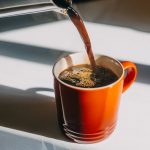 Coffee being poured into an orange mug on a white countertop.